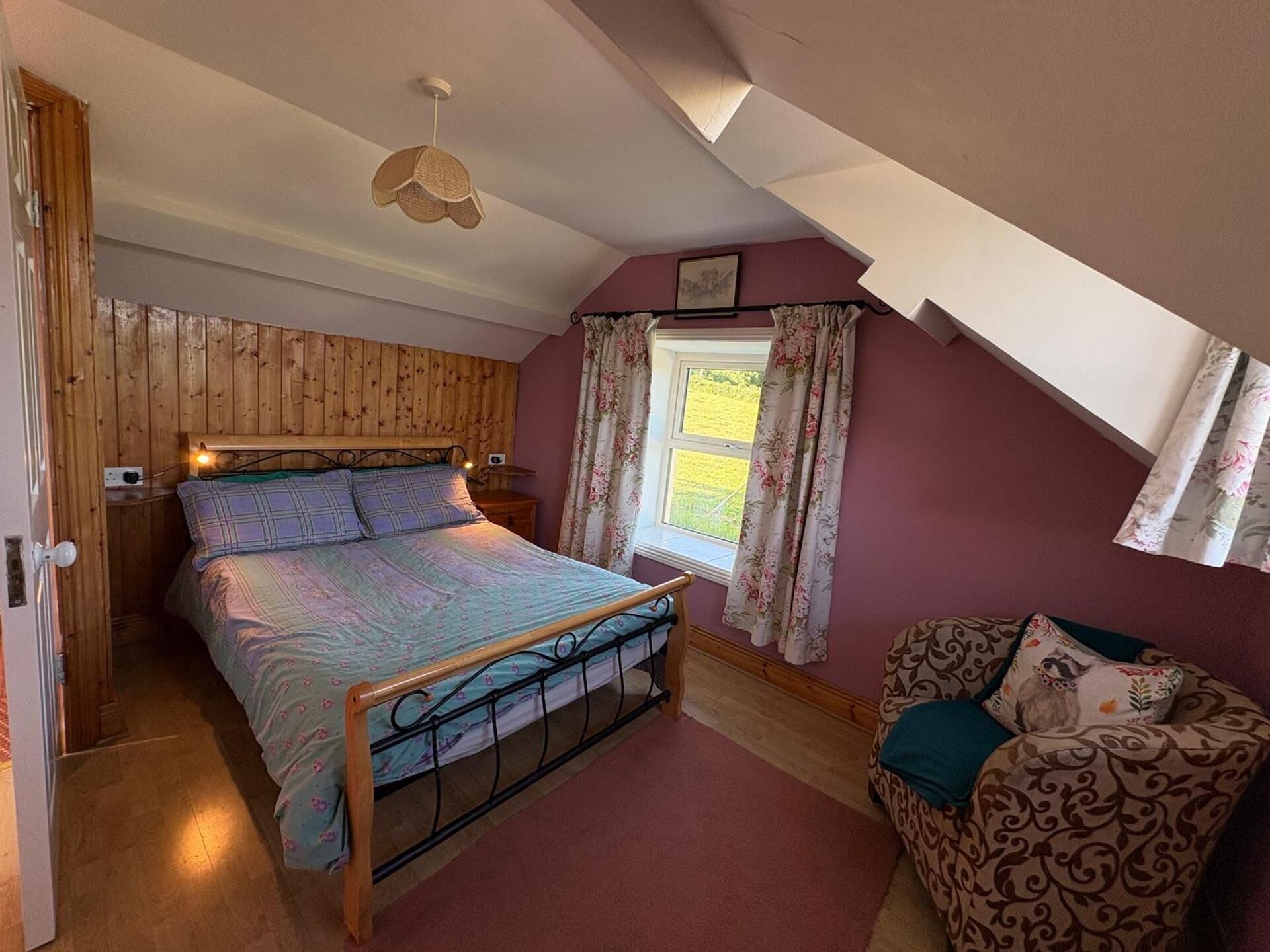 A bedroom with a wooden headboard, a bed with a patterned bedspread, a nightstand, a desk, a chair with a floral cushion, a window with curtains, and a hanging light fixture.