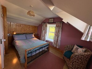A bedroom with a wooden headboard, a bed with a patterned bedspread, a nightstand, a desk, a chair with a floral cushion, a window with curtains, and a hanging light fixture.