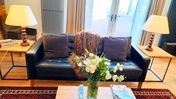 A modern living room with a black leather sofa, two lamps, a vase with flowers, and a remote control on a wooden coffee table.