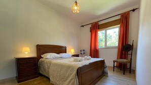 Un dormitorio con una cama de madera, dos lámparas de noche, un aparador de madera, un ventanal con cortinas y una silla de madera.