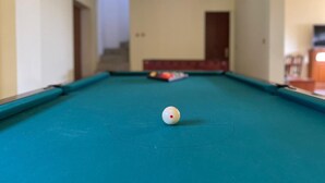 Una mesa de billar con una bola blanca en el centro, un punto rojo sobre la mesa y un juego de tacos de billar al fondo.
