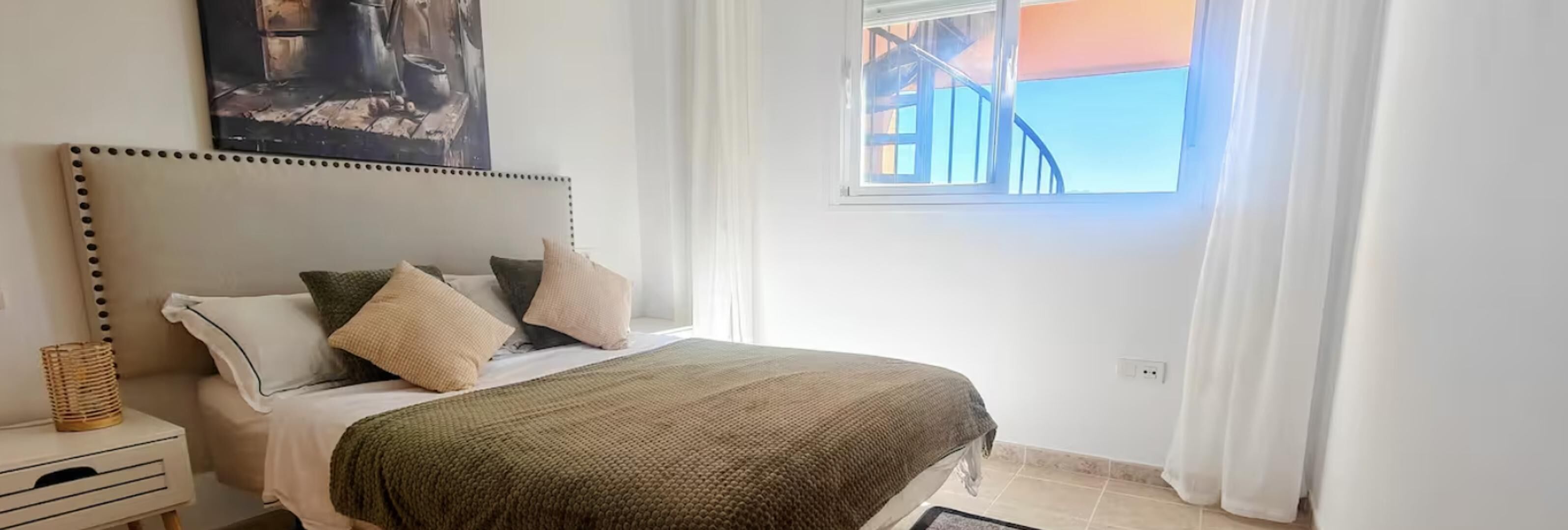 A bedroom with a bed, pillows, a bedside table, a window with curtains, and a painting on the wall.