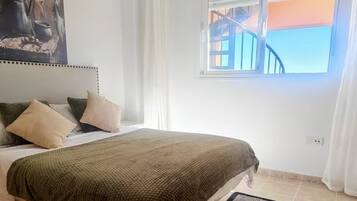 A bedroom with a bed, pillows, a bedside table, a window with curtains, and a painting on the wall.