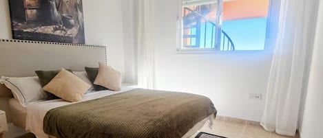 A bedroom with a bed, pillows, a bedside table, a window with curtains, and a painting on the wall.