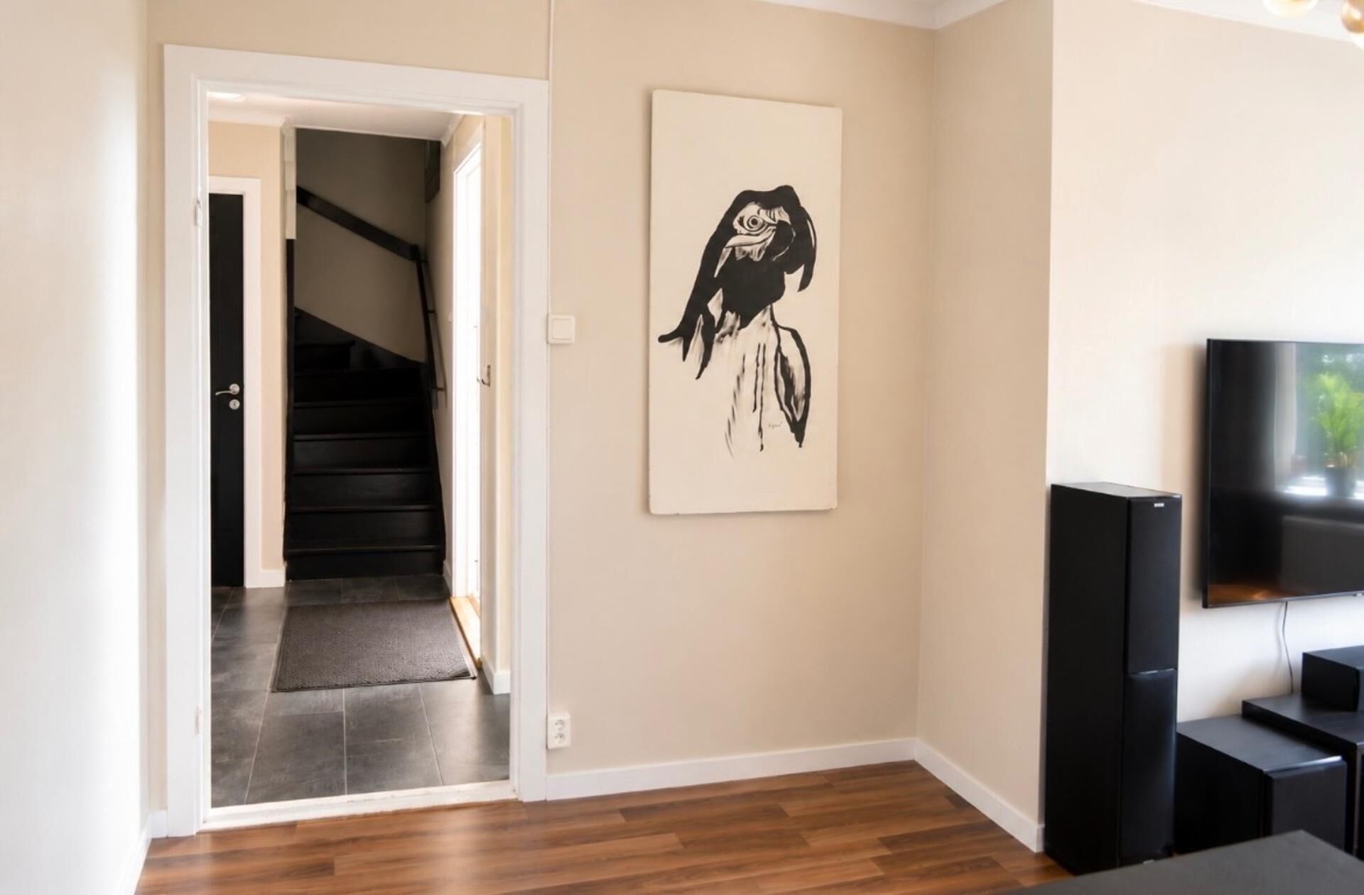 A modern living room with a staircase leading to a dark-toned hallway, a black and white artwork on the wall, and a flat-screen TV on a stand.