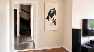 A modern living room with a staircase leading to a dark-toned hallway, a black and white artwork on the wall, and a flat-screen TV on a stand.