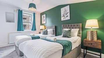 A double bed with a grey headboard and white bedding, a bedside table with a lamp, and a window with curtains.