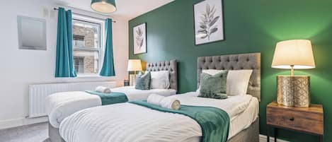 Una cama doble con cabecera gris y ropa de cama blanca, una mesita de noche con lámpara, y una ventana con cortinas.