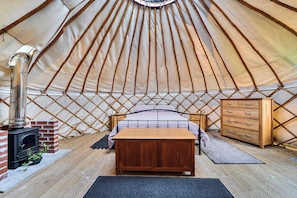 A cozy interior of a yurt with a bed, a dresser, and a wood-burning stove. - Yurt Sleeps 2 - Bbq & Fire Pit - Dogs Welcome (Bodmin)