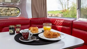 Une table avec des croissants, une tasse de café et un verre de jus d’orange dans un wagon de train aux sièges en cuir rouge, avec vue sur la verdure environnante.