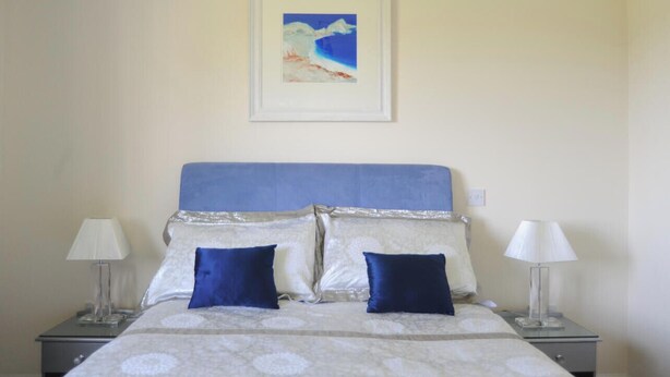 A neatly made bed with a blue headboard, two pillows, and a white blanket. There are two bedside lamps on either side of the bed. - Spacious Bungalow Sleeps 10 (Kidwelly)