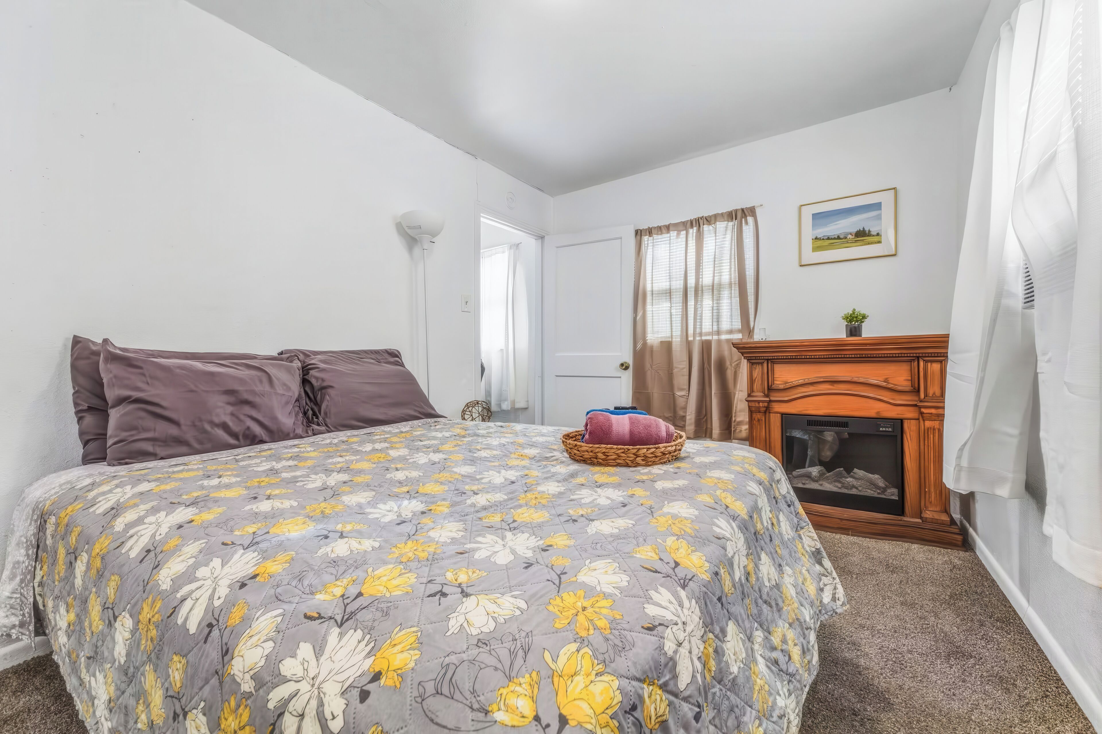 A bedroom with a bed, pillows, a bedside table with a lamp, a fireplace, and a framed picture on the wall.