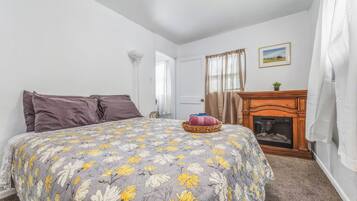 A bedroom with a bed, pillows, a bedside table with a lamp, a fireplace, and a framed picture on the wall.