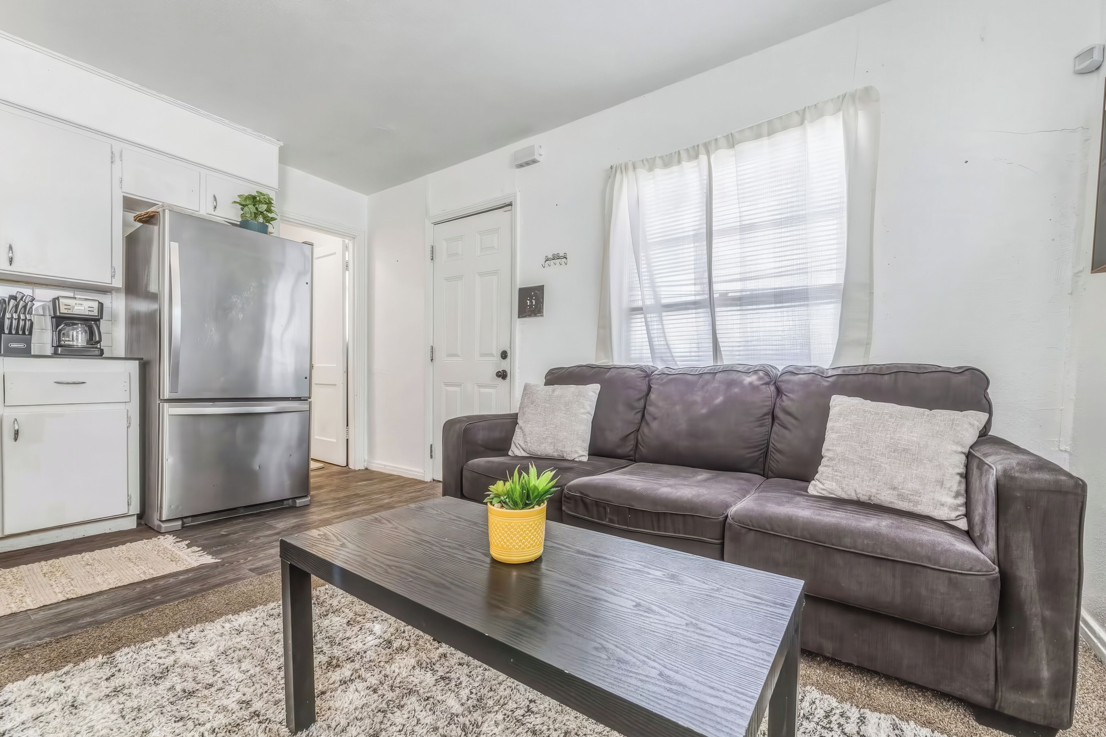 A modern living room with a grey sofa, a coffee table with a plant, and a kitchen area with a refrigerator and cabinets.