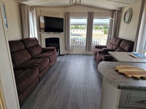 A living room with a flat-screen TV, two sofas, a small table, and a kitchen area with a wooden countertop. - Caravan Sleeps 6 With Parking (Great Yarmouth)