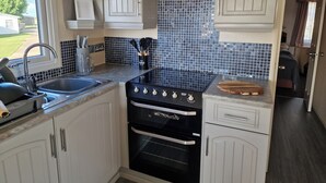 A compact kitchen with white cabinets, a black stove, a sink, and a backsplash with blue tiles. - Caravan Sleeps 6 With Parking (Great Yarmouth)