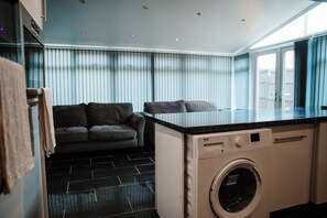 A compact laundry area with a washing machine, a small sofa, and a window with blinds. - 2 Bed House Sleeps 4 -private Garden -free Parking (Bury)