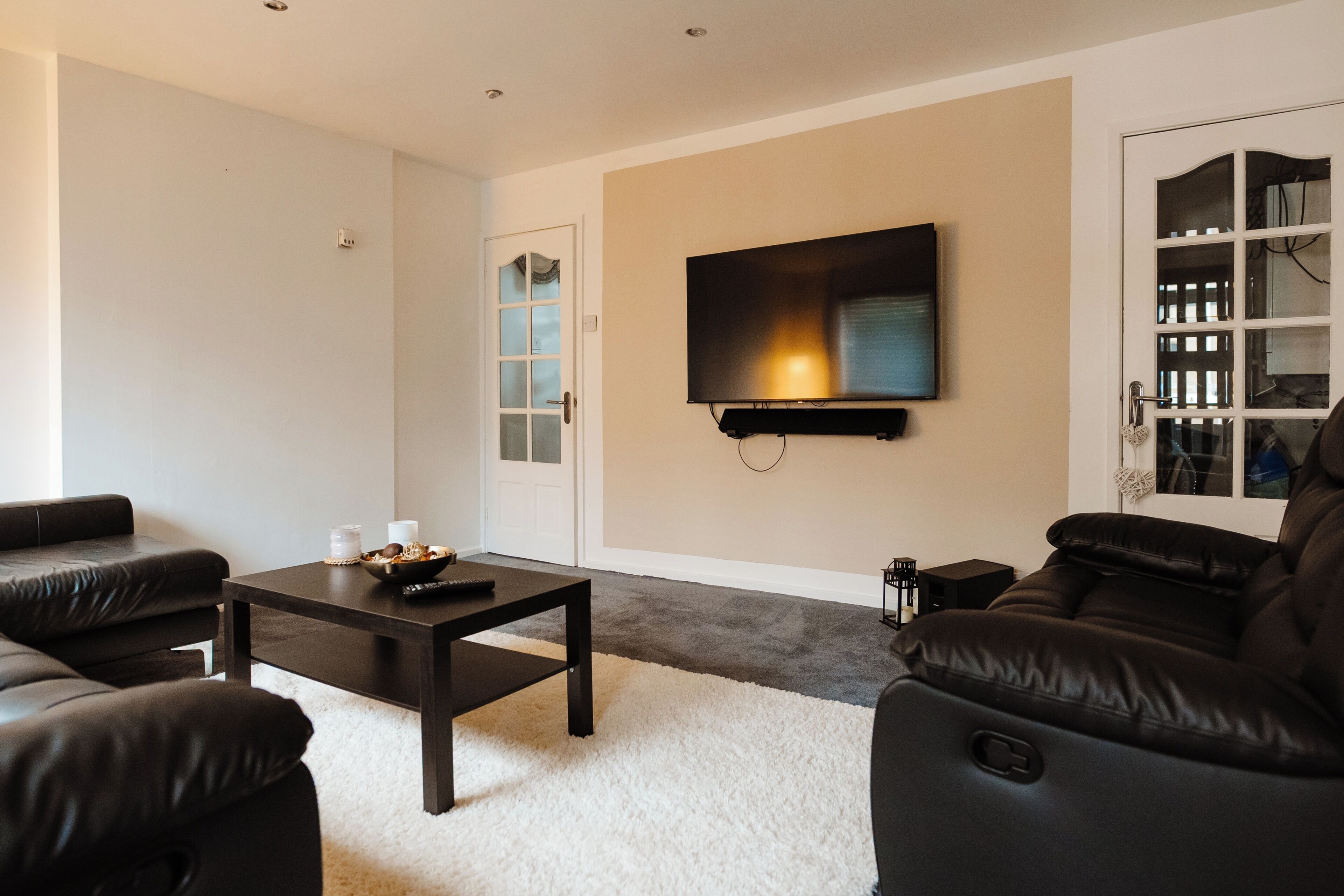Un salon moderne avec un téléviseur à écran plat fixé au mur, une table basse en bois foncé et des canapés en cuir noir.