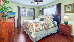 A hotel room with a bed, a ceiling fan, a TV, a plant, a dresser, a nightstand, and a window with curtains. - NEW! Beachfront 2BR + Pool + Walk to Beach (Indian Rocks Beach)