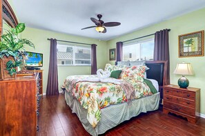 Ein Hotelzimmer mit einem Bett, einem Deckenventilator, einem Fernseher, einer Pflanze, einem Schminktisch, einem Nachttisch und einem Fenster mit Vorhängen.