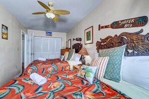 A bedroom with a bed, a ceiling fan, a wooden headboard, and a surfboard decoration. - NEW! Beachfront 2BR + Pool + Walk to Beach (Indian Rocks Beach)