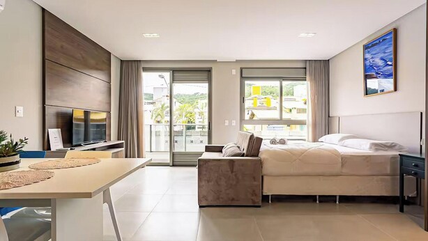 A modern hotel room with a bed, a desk, a TV, and a view of the city through large windows. - Amazing Studio 5min Walk to the Beach Elu109 (Florianópolis)