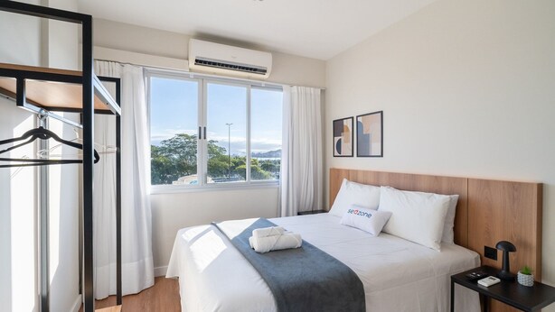 A hotel room with a bed, a nightstand, a window with curtains, and a view of the outdoors. - Fully Equipped apt in Downtown Floripa - Ejo0306 (Florianópolis)
