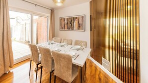 A dining area with a table set for six, a chandelier, a large abstract painting, and a view of a brick wall and garden through a sliding door. - Spacious 5-bedroom House in Berkshire - Perfect Family Retreat (Reading)
