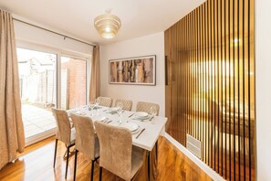 A dining area with a table set for six, a chandelier, a large abstract painting, and a view of a brick wall and garden through a sliding door. - Spacious 5-bedroom House in Berkshire - Perfect Family Retreat (Reading)