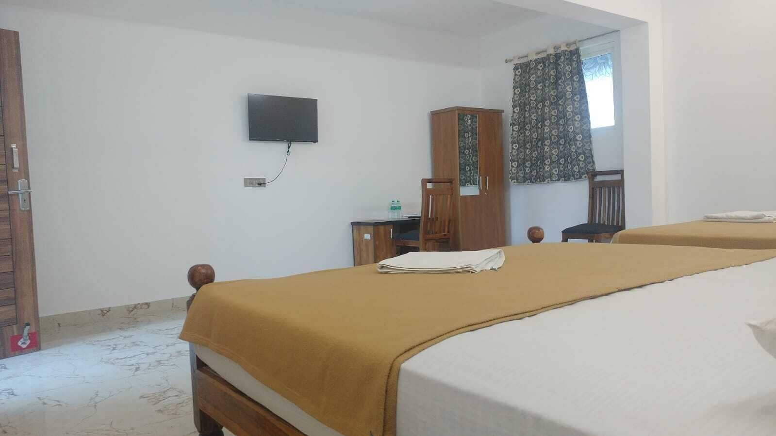 Habitación de hotel con dos camas, un televisor montado en la pared, un armario de madera y una ventana con cortinas.