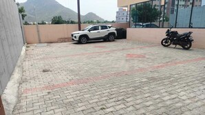 A paved outdoor area with a white SUV parked, a motorcycle, and a building with a glass facade in the background. - SJS Residency by Orange Hotels (Tiruvannamalai)