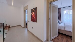 Quarto de hotel com cama, janela com cortinas, quadro na parede e corredor que dá para outro quarto.
