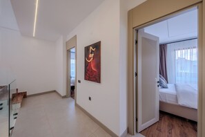 A hotel room with a bed, a window with curtains, a painting on the wall, and a hallway leading to another room.