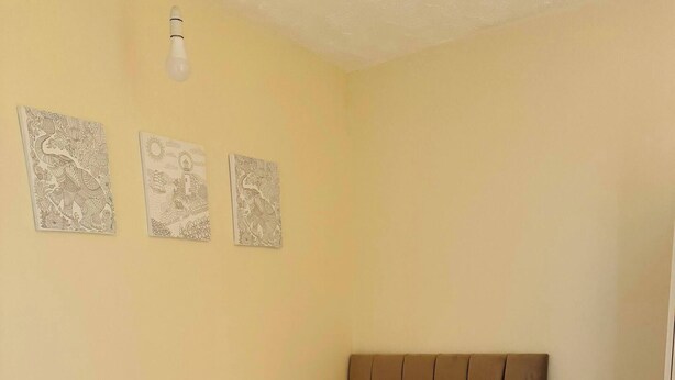 Cama de casal com cabeceira marrom, roupa de cama branca e mesa de cabeceira com abajur. Há três quadros na parede acima da cama.
