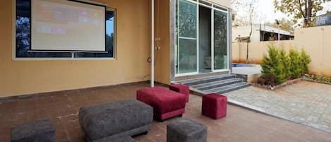 Un espacio exterior con mesas, una pantalla de proyector, la entrada de un edificio moderno y un patio empedrado.