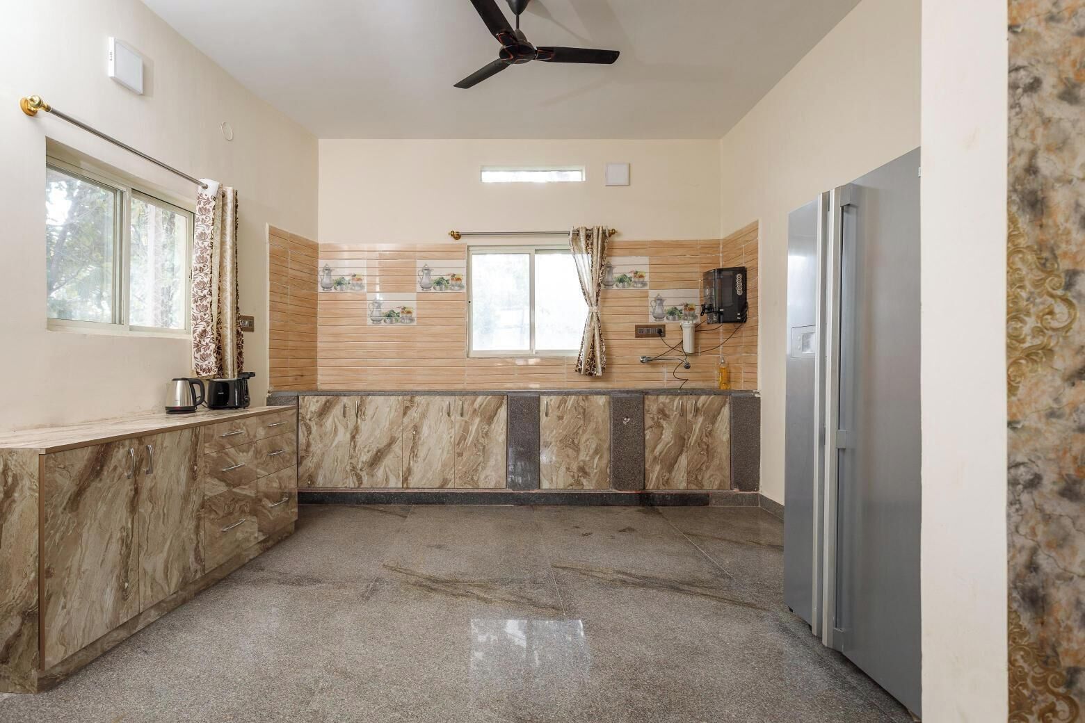 A kitchen with marble countertops, a ceiling fan, a refrigerator, and a window with curtains.