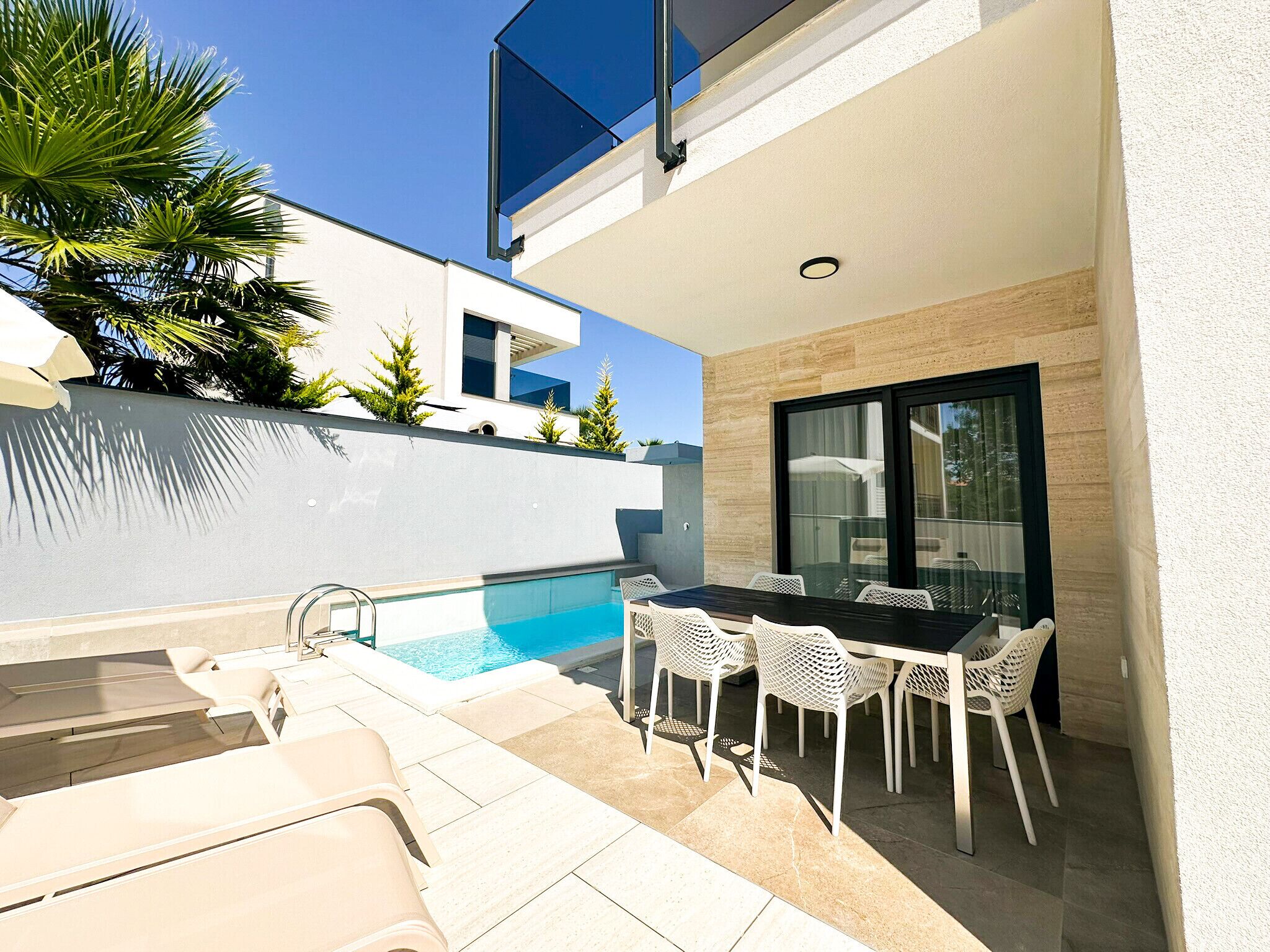 Un área de piscina exterior moderna con una mesa y sillas para comer, rodeada por un muro blanco y un puerta de cristal.