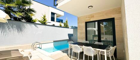 A modern outdoor pool area with a dining table and chairs, surrounded by a white wall and a glass door.