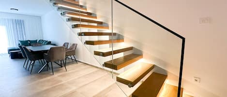 A modern staircase with glass railings and wooden steps, leading to a dining area with a table and chairs.