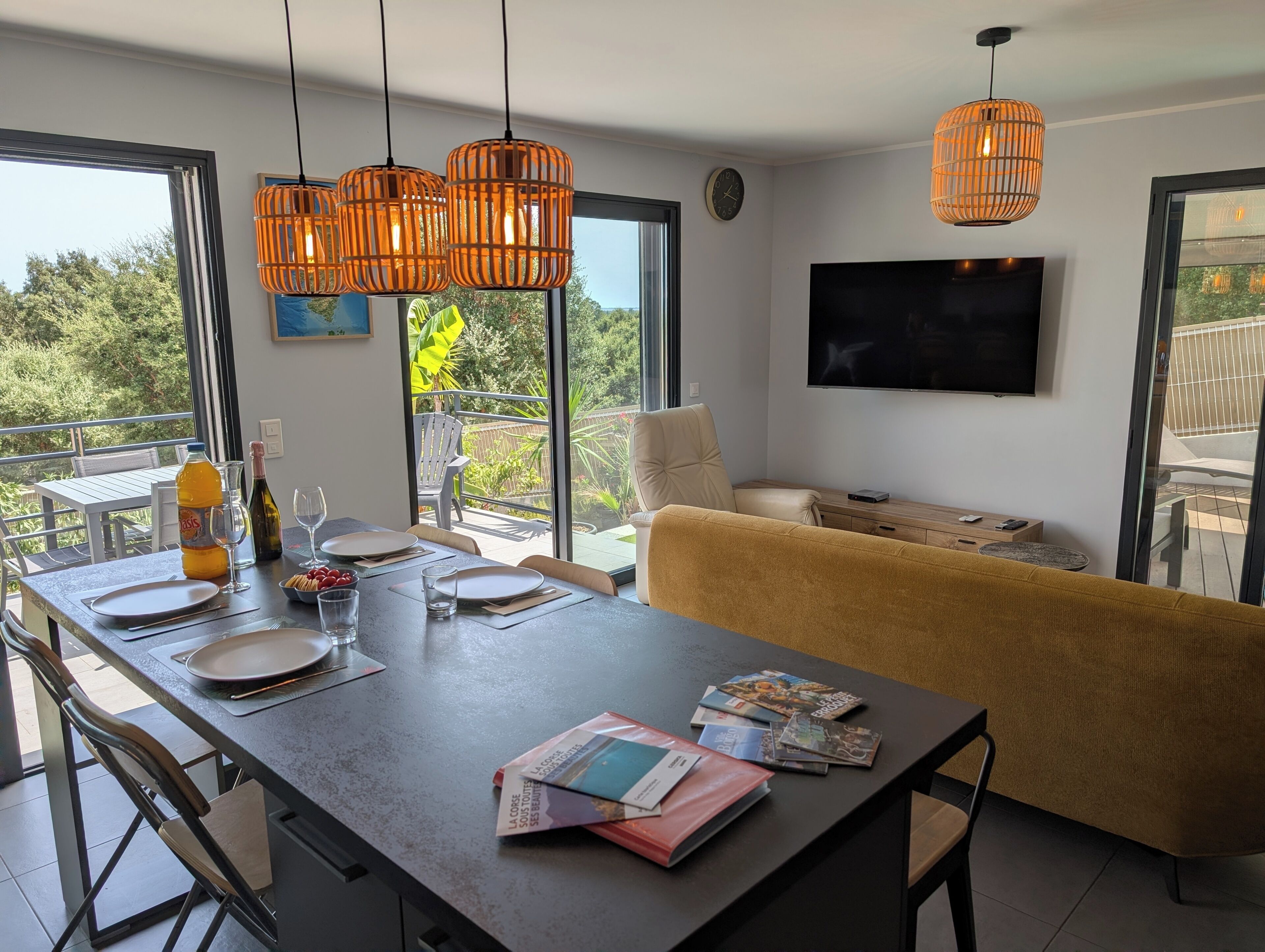 A modern living space with a dining area, sofa, TV, and large windows offering a view of greenery.