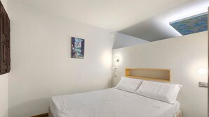 Une chambre d’hôtel avec un lit simple, une tête de lit en bois, une lampe fixée au mur et un tableau représentant un paysage de montagne.