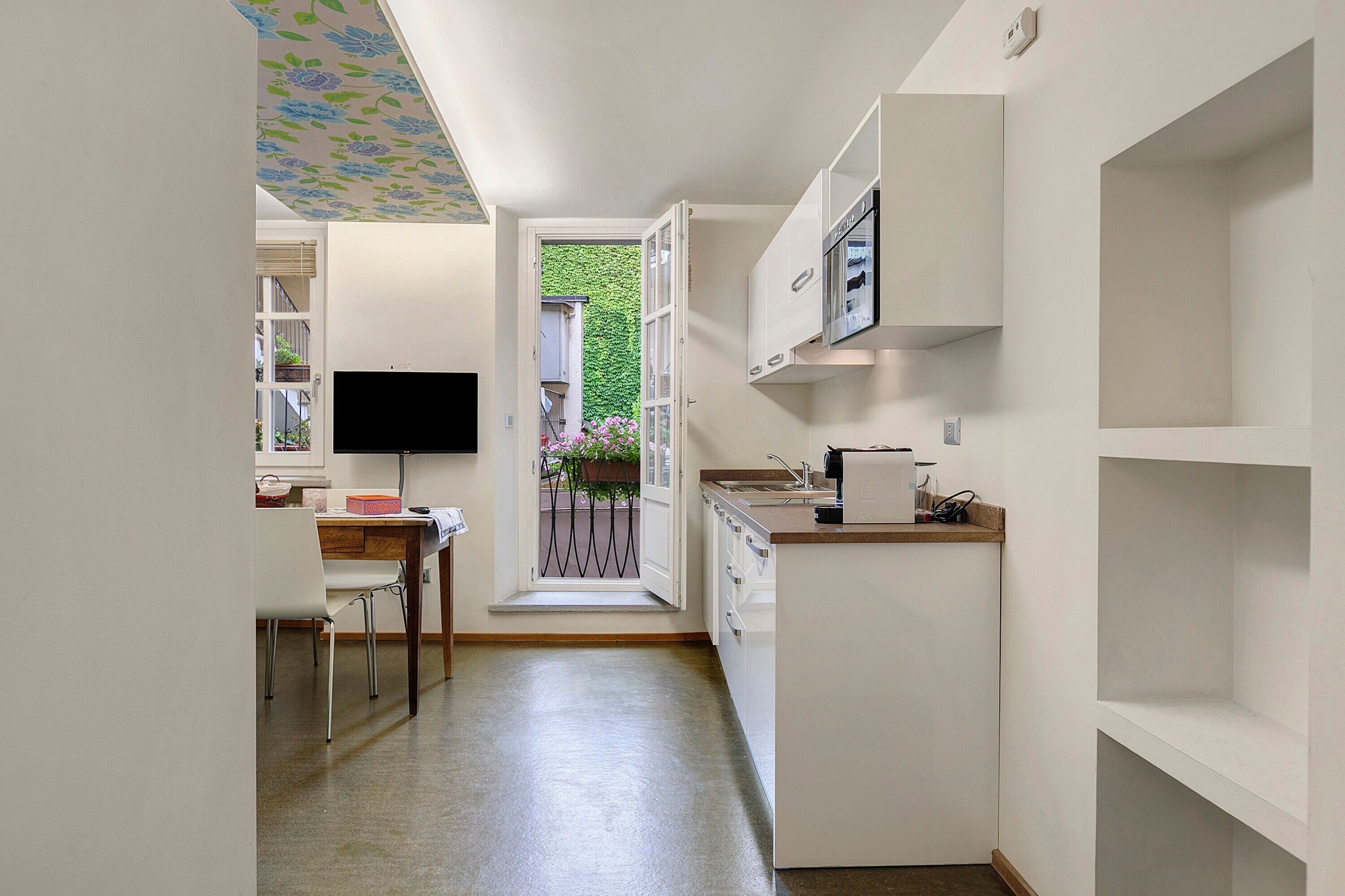 A narrow kitchen with a dining area, a television, and a view of a balcony with plants.