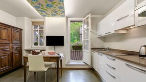 Une cuisine avec des meubles blancs, une table à manger en bois, un téléviseur et une vue sur un jardin par une porte ouverte.