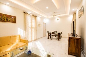 A living room with a sofa, a glass coffee table, a dining table with chairs, a television on a wooden stand, and a painting on the wall. - Marigold by Red Olive 3bhk Nr Fortis 20min DLF (Gurugram)