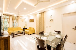 A living room with a yellow sofa, a glass dining table set for four, and a ceiling fan. - Marigold by Red Olive 3bhk Nr Fortis 20min DLF (Gurugram)