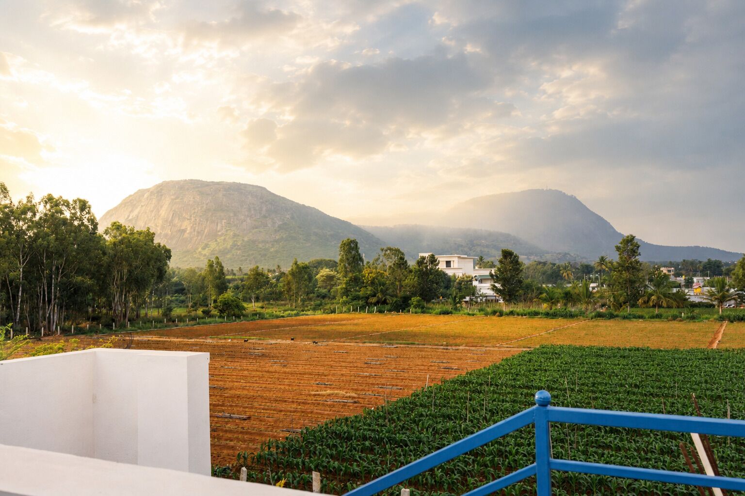 A rural landscape with fields, a mountain, and a white building with a blue railing in the foreground.