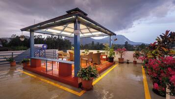 A covered outdoor seating area with chairs and tables, surrounded by potted plants and a view of a mountain.