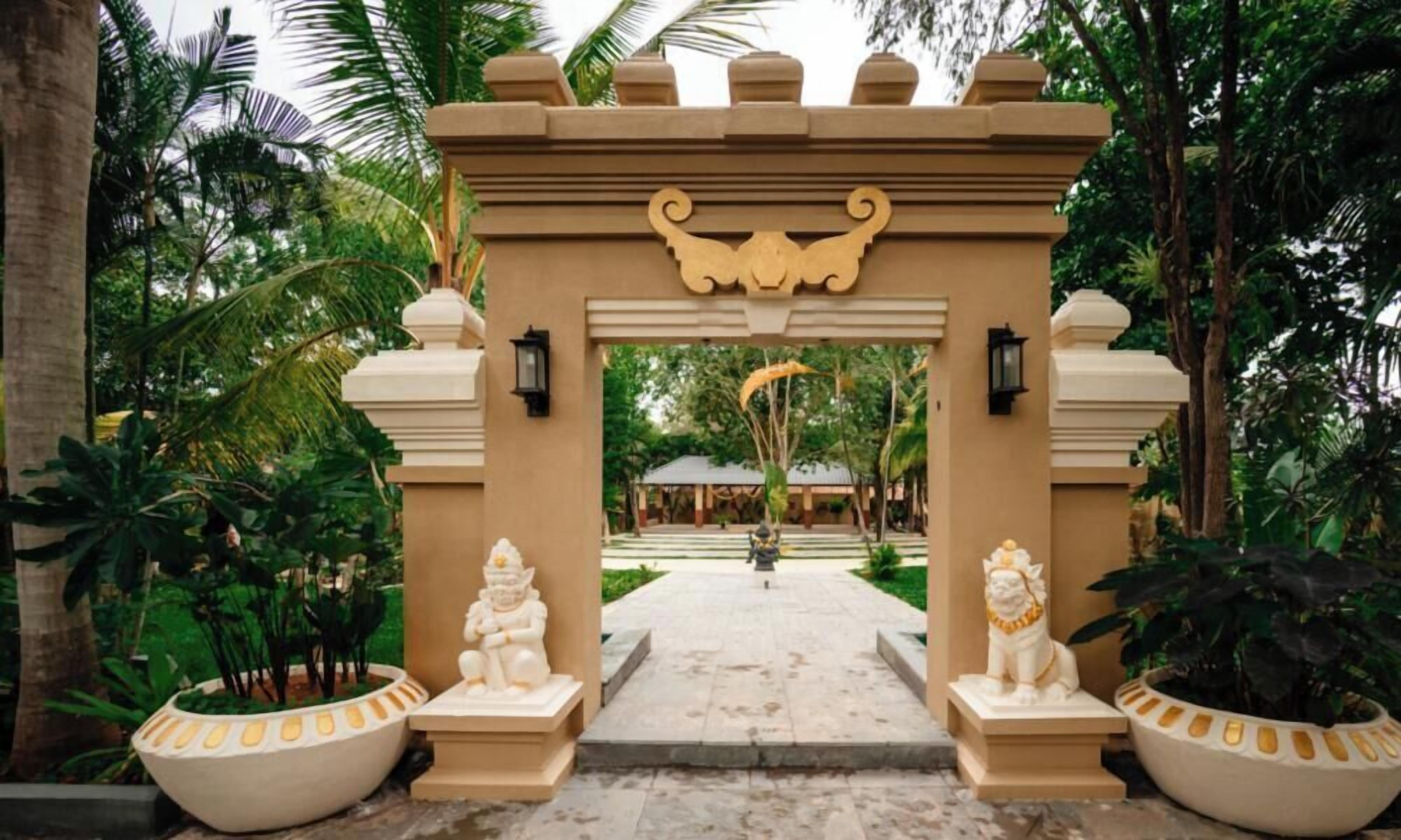 Uma entrada tropical com um arco decorativo, duas estátuas e um caminho que leva a um edifício.