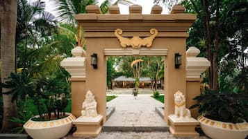 Uma entrada tropical com um arco decorativo, duas estátuas e um caminho que leva a um edifício.