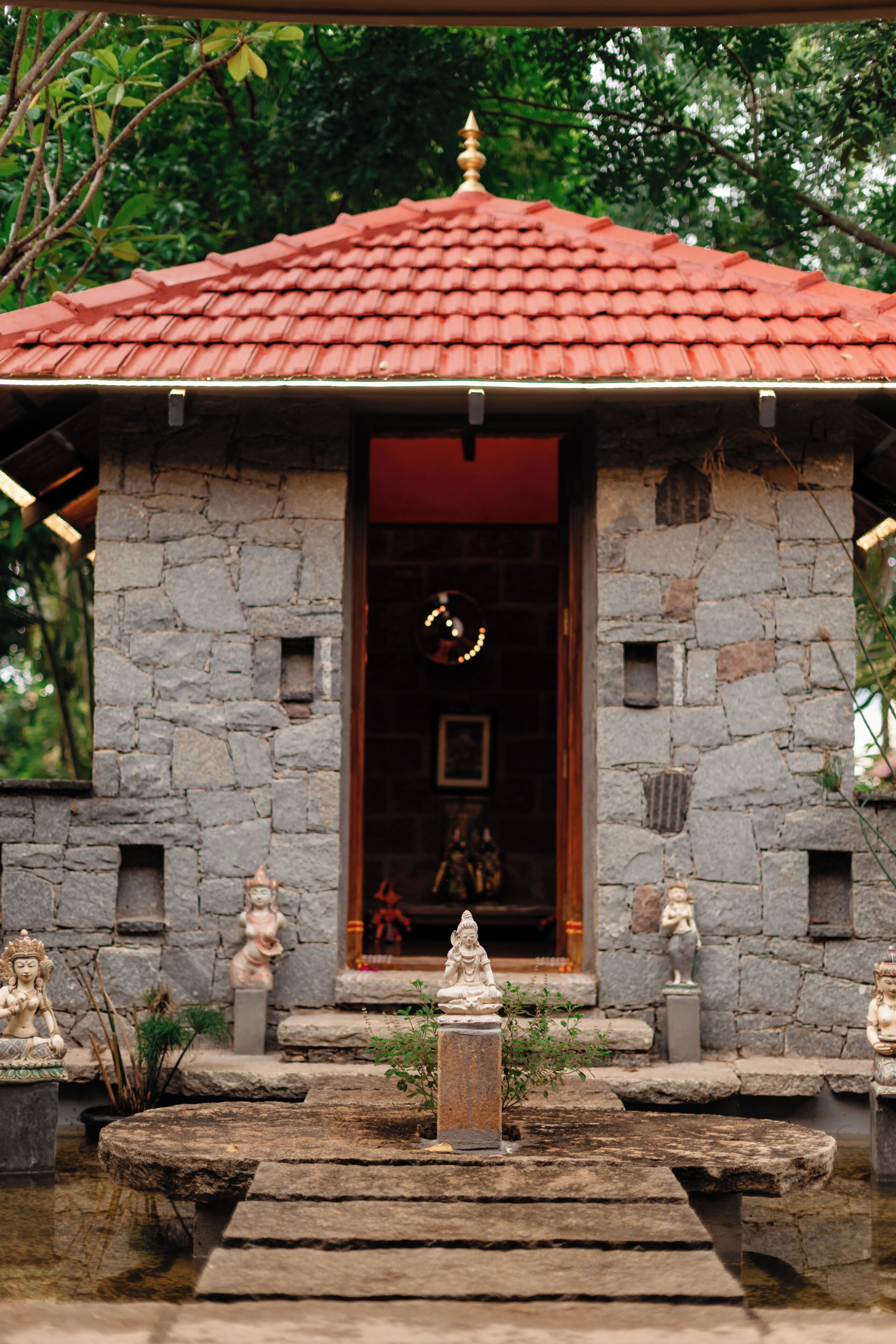 A stone structure with a red-tiled roof, a central doorway, and statues around a water feature.